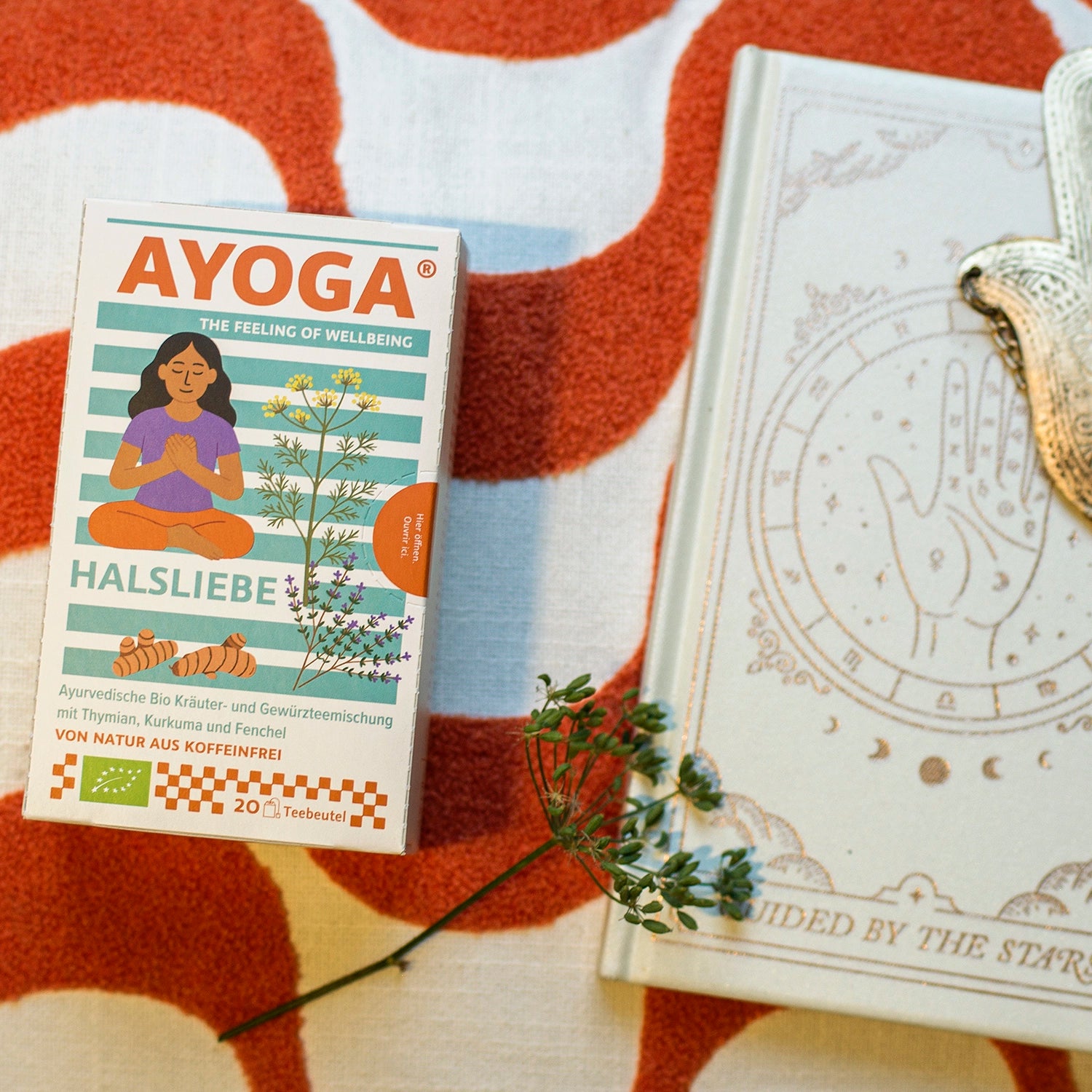 A box of 'AYOGA Halsliebe' on a red patterned carpet, next to a book.